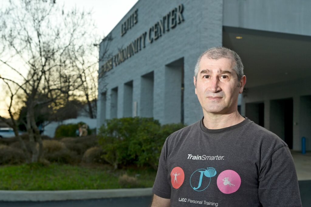 A well-lit man with very short hair and wearing a t-shirt stands in front of the Levite Jewish Community Center at dusk