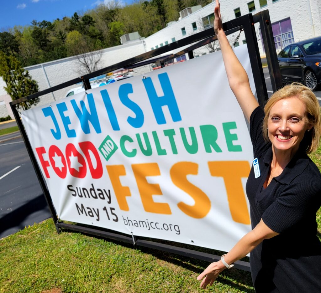 A blonde woman wearing black stands in a playful pose with her arms stretched upward and downward to frame a large banner that's advertising the "Jewish Food and Culture Fest"