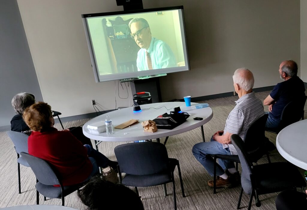 Four people in a darkened room sit around a round table watch a movie on a large TV screen