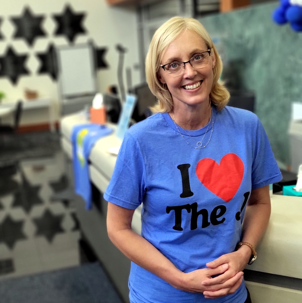 A woman in an "I love The J" t-shirt leans on the Welcome Desk as she poses in the LJCC lobby