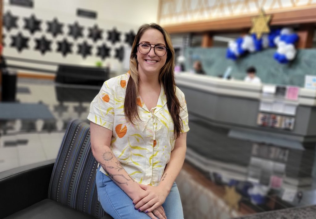 A woman in a light-colored patterned untucked button-down shirt perches on the arm of a chair in the LJCC lobby.