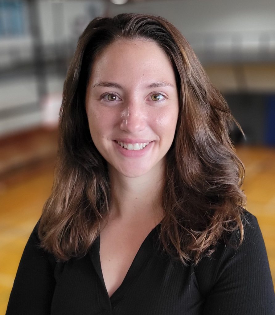 A headshot of a woman with long hair and wearing a black dress with a gymnasium floor in the background