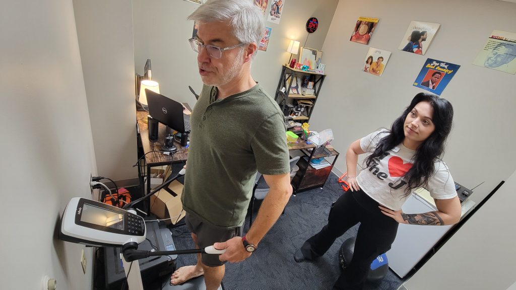 A man in an office stands on an scale-like device while a woman stands behind him monitoring his results