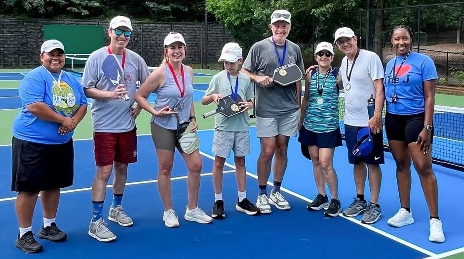 Eight pickleball players pose in a line on an outdoor court after winning various elements in a pickleball tournament