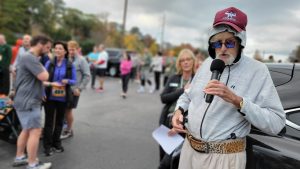 A man wearing a cap over his hoodie and sporting a leopard-patterned lifting belt sings into a microphone in a parking lot