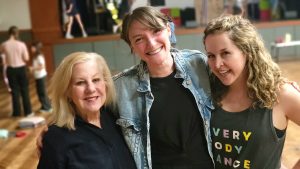 Three women stand together and smile broadly as Suessical Jr. cast members rehearse in the background.