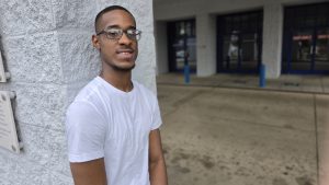 A young man in a white t-shirt poses by a pillar in front of The J's entrance