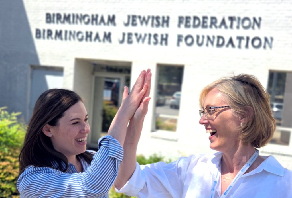 Two women high-five in front of a white building with "Birmingham Jewish Federation" and "Birmingham Jewish Foundation" written on it