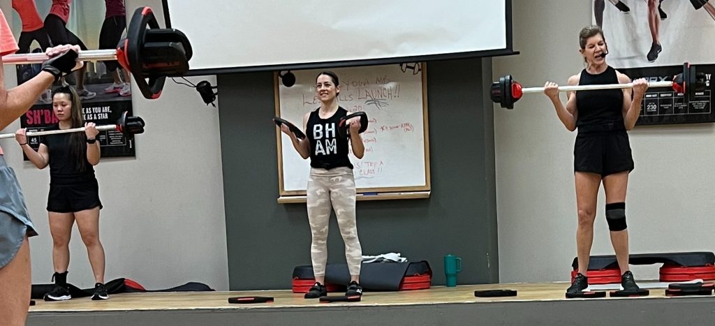Three women on a stage lift weights while leading an intense group fitness workout session