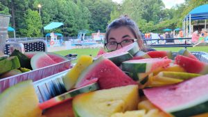 A woman in glasses peeks over the top of a pan of sliced watermelon ; the outdoor pool is visible in the background
