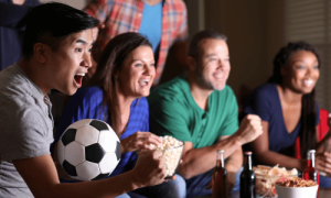 Four people sitting on a couch watch a soccer game while one holds a soccer ball