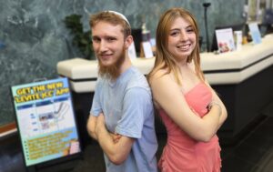 A man and a woman pose back-to-back with arms folded in front of The J's Welcome Desk